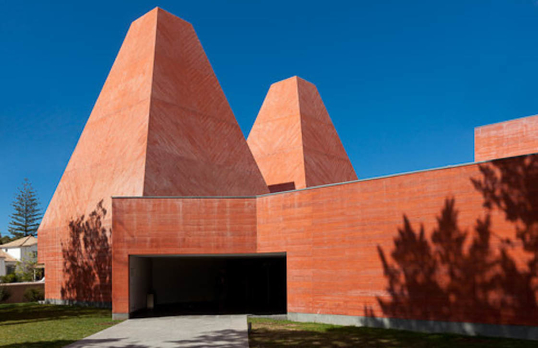An orange pink clay coloured building with two pyramids by Eduardo Souto de Moura