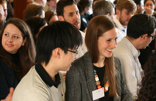 Two smiling young adults, one with short black hair and one with long ginger hair, laughing while surrounded by a seated crowded