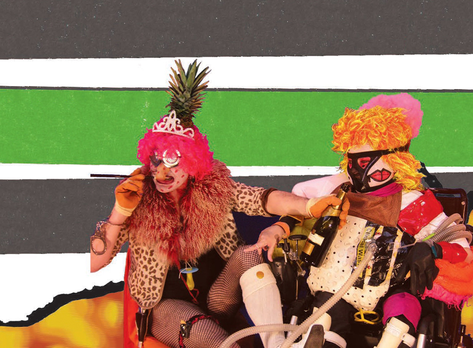 Two disabled people in colourful fancy dress and ridiculous headwear with a bottle of champagne. They seem to be bursting with energy, leaping up in front of the two green strips from the cover of UK accessibility Part M documents.