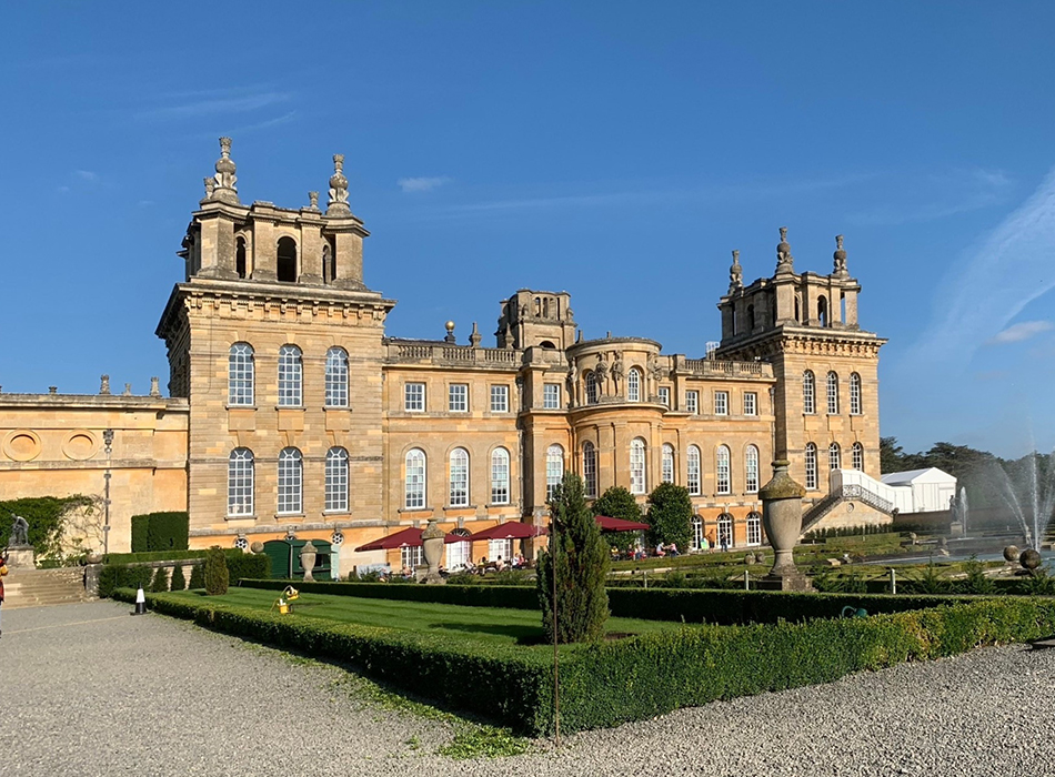 Blenheim Palace - a sandstone building in baroque style with arched windows and decorative gardens. Pictured in sunlight with blue sky.