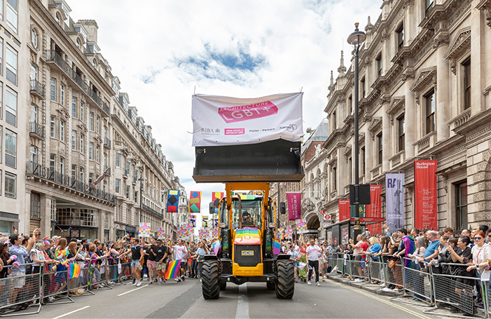 A rainbow painted digger with an Architecture LGBT+ banner and pride parade marchers behind it, with people waving rainbow flags on both sides of the road