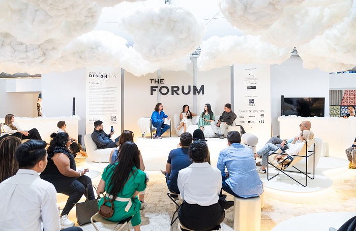 Selection of people gathered around a stage set up for four particpants. There are faux clouds covering the ceiling and the word FORUM in large black letters hangs over the stage.