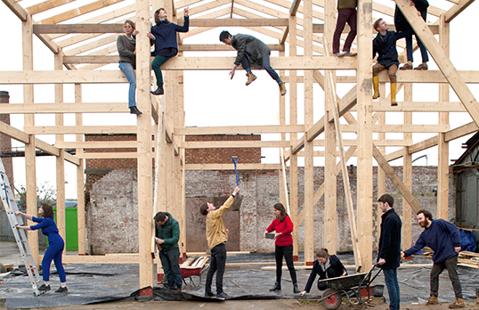 Various adults in street clothes working on a timber frame building