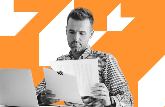 A man reading a piece of paper whilst sat by a laptop with an orange background 