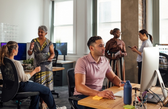 A group of coworkers of a variety of races in an office. They are working and some are conversing.