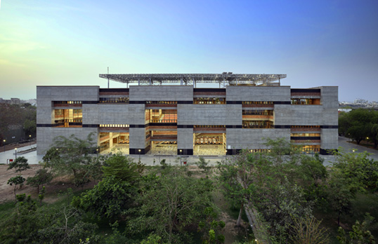 A straight on view of Ahmedabad University centre, a modern building with large open sections and windows lit up.