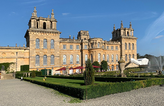 Blenheim Palace - a sandstone building in baroque style with arched windows and decorative gardens. Pictured in sunlight with blue sky.