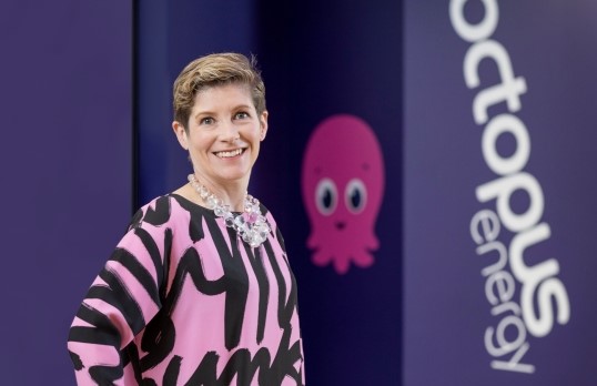 Woman with short hair wearing pink and black smiling at camera  with ocotpus energy purple branding background and the iconic pink Constantine octopus mascot on the wall