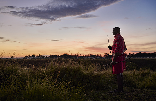 Maasai man wearing red holding a bow and arrow standing in a grassy plain at sunset