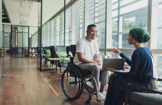 Two adults sitting in a modern office space, one with a head covering, holding a laptop and facing another who is sitting in a wheelchair