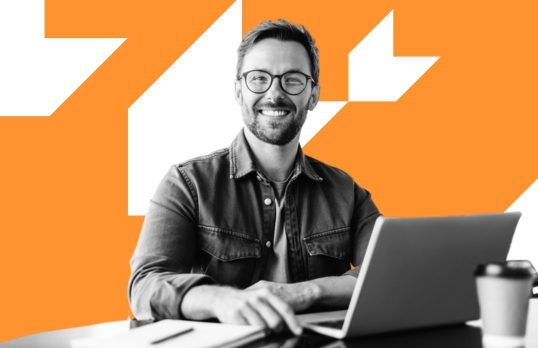 Man wearing glasses working at laptop smiling to camera with orange background