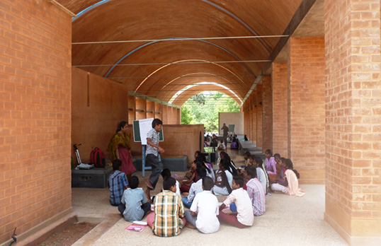 People gathered to learn inside the Sharanam Centre. The building is red brick with an arched ceiling that at the far end opens out into greenery.