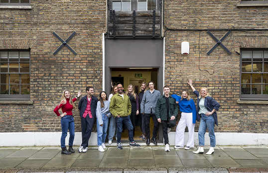 Team of 11 architects smiling at camera stood in the doorway of a brown brick building with cross structural supports.