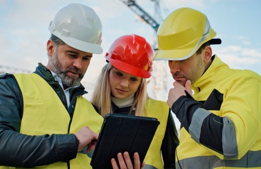 Three architects on a building site wearing high visibility jackets and looking at a digital tablet 