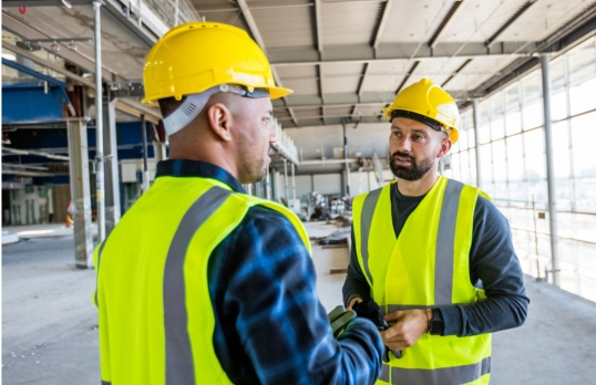 Two men wearing hard hats ad hiviz jackets talking on a building site 