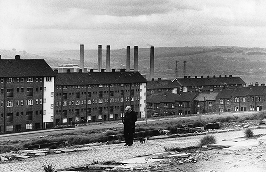 Black and white photo of person standing alone in front of row of mill buildings with chimneys in skyline behind.