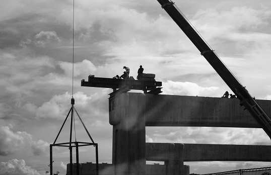 Black and white image of workers on a high beam with cranes overhead and cloudy sky behind.