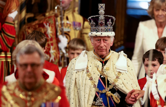 King Charles III wearing crown and full royal garb processing into Westminster flanked by officials.