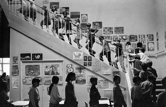 Black and white photo of school children walking up a staircase with drawings on the wall