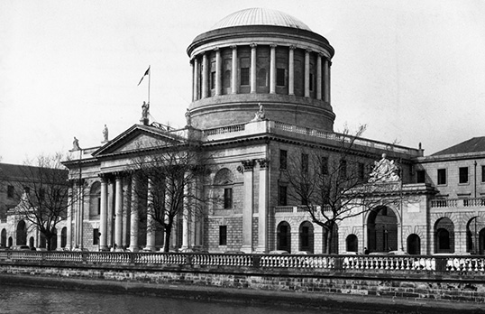 Black and white photograph of a neoclassical building along the River Liffey in Dublin with square base, columned entrance and columned dome tower