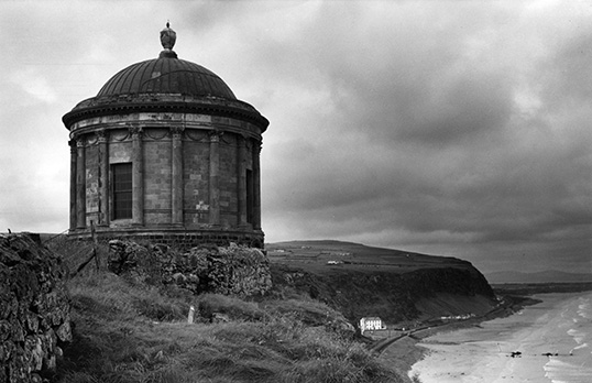 Black and white photograph of a neoclassical temple building with columns and rounded roof overlooking a small cliff and beach