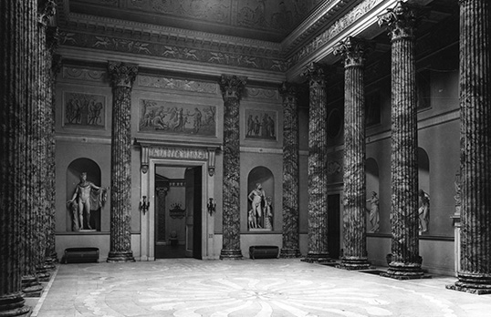 Black and white photograph of a large hall interior with decorated neoclassical columns and tile mosaics on the floor and walls