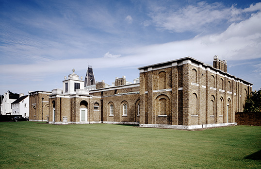 Coloured photograph of Dulwich Picture Gallery, a neoclassical brick building with white trim and a small domed tower, surrounded by grass