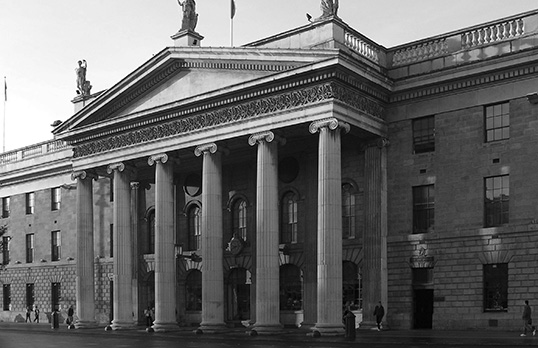 Black and white photograph of the General Post Office in Dublin, a multi-storey stone building with neoclassical columned front and triangular feature on a flat roof