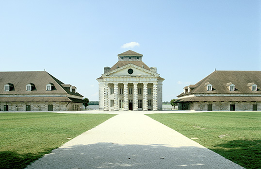 Coloured photograph of a central building with neoclassical columns and two shorter side buildings with gable roof, grass and a long white path in the foreground