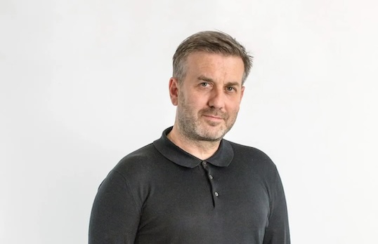 Man wearing black polo short with swept back hair and fashion stubble standing against a what wall and smiling at camera