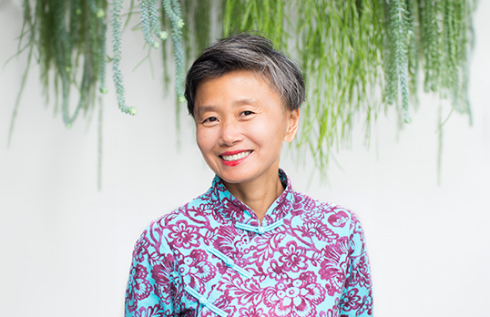 Woman with short grey hair and wearing blue and purple patterened top standing in front to white wall with grasses hanging vertically down
