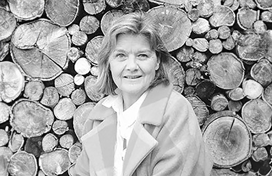 Black and white photograph of woman wearing thick coat in front of a pile of logs