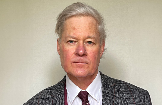 Older man with white swept over hair and wearing a shirt and tie with a patterned blazer in front of a cream wall