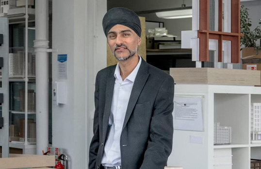 Man with facial hair and wearing pale blue shirt with a dark blue suit standing in office full of storage and posts