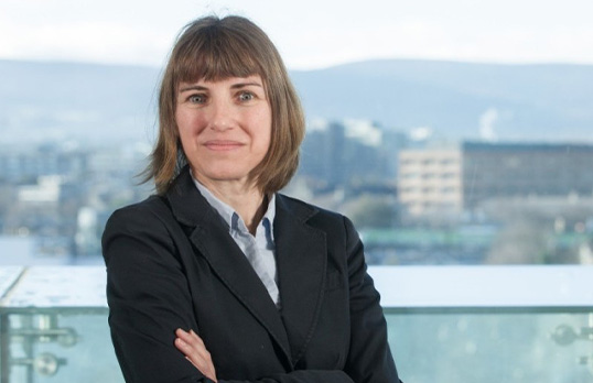 Woman with brown hair and wearing pale blue shirt and dark grey suit with her arms folded in front of a skyline background.