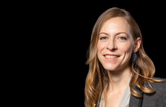Woman with long brown wavy hair and wearing grey against a black background