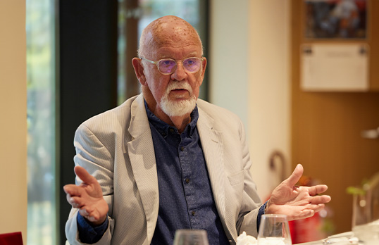 Older man with white goatee beard wearing dark blue shirt and cream suit with their hands outstretched sat at a table in an office environment.
