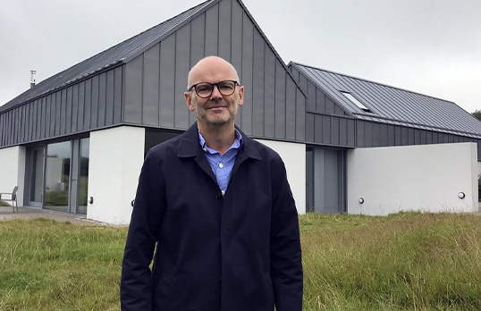 Bald man with glasses wearing a blue shirt and shacket in front of a barn style building surrounded by grass.