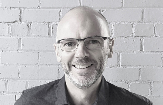 Bald man with glasses, smiling at camera, white brick wall behind