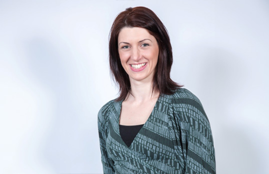 Woman with long brown hair and wearing a patterned blue and black top and smiling at camera
