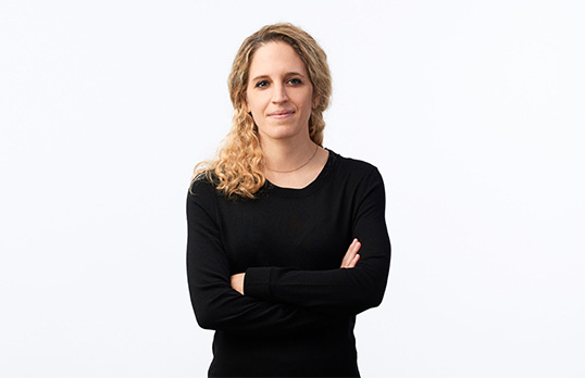 Woman with wavy blond hair and wearing a black top with her arms folded standing against a white wall.