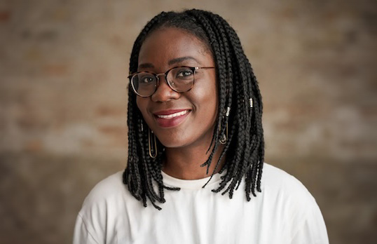 Woman with black braids and glasses wearing a white tee in a brown brick room