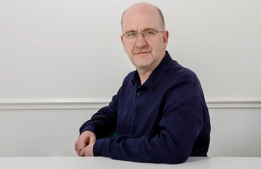 Bald man with glasses side on to camera wearing a dark blue shirt and sitting behind a table.