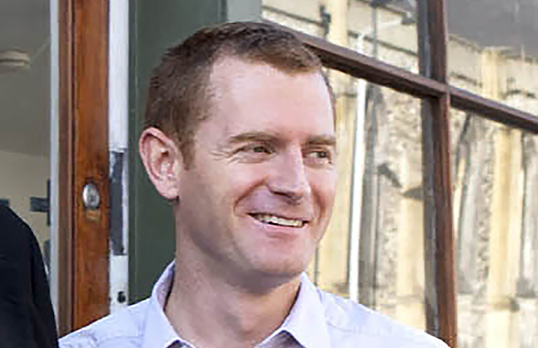 Man with short brown hair wearing pale blue shirt standing in front of a shop front.