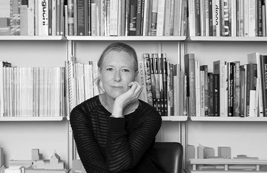 Woman in black with head resting on hand sitting in front of bookcase.