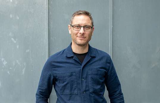 Man with short hair and glasses wearing a blue shirt over black tee and standing in front of zinc cladding