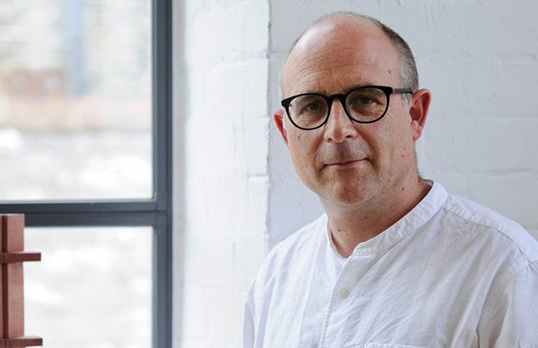 Bald man with glasses wearing white grandad-collar shirt standing next to a window