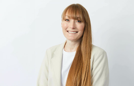 Woman with very long strawberry blond hair and wearing a cream suit standing in a white room.