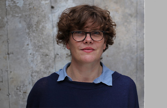 Woman with short curly hair and fringe wearing glasses and a blue shirt and jumper standing in front of a rough concrete wall