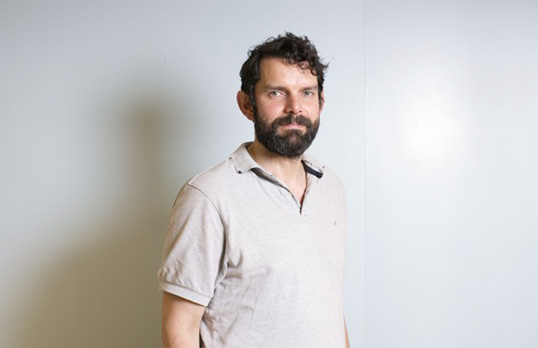 Man with dark hair and beard wearing an off-white polo short and standing in a white room.